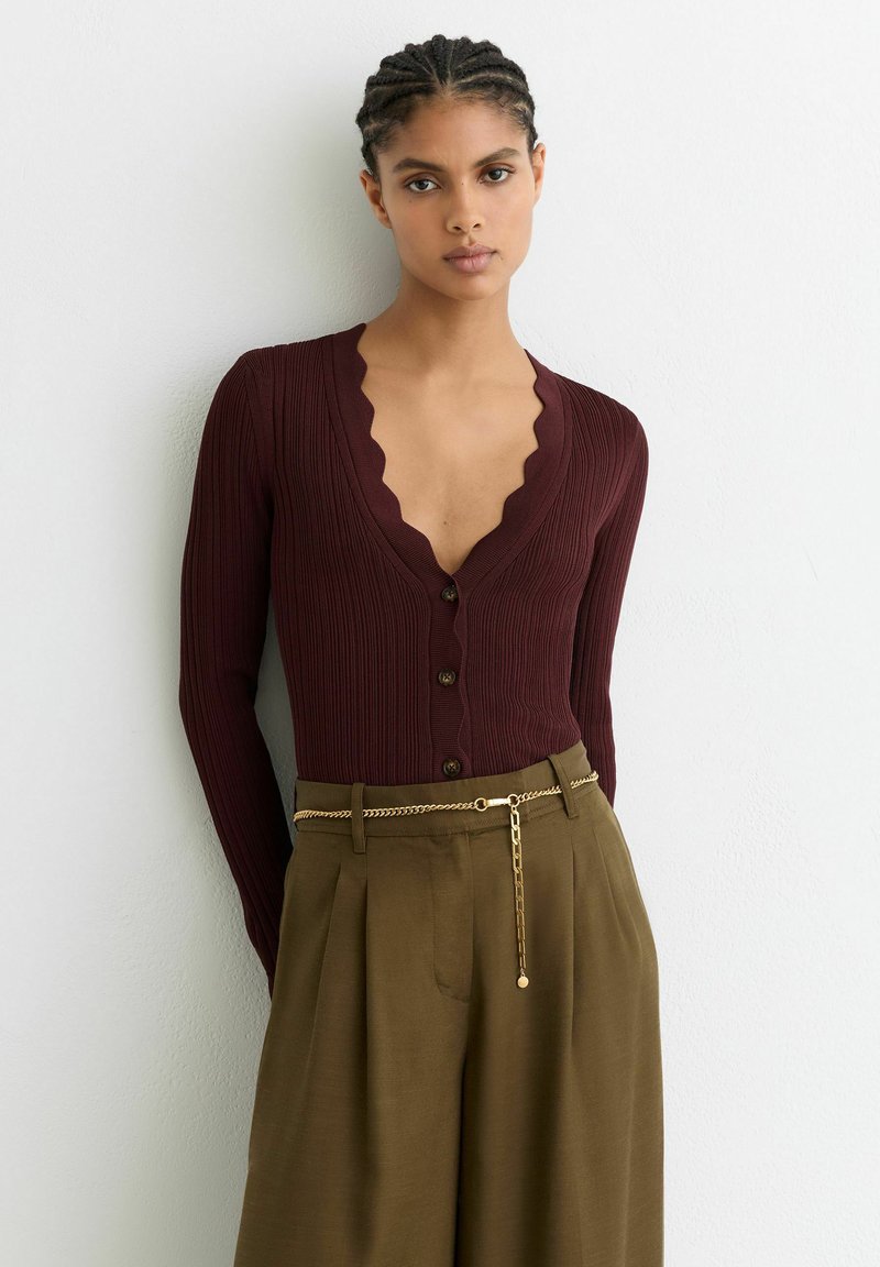 Bordeauxrode geribbelde cardigan met een geschulpte halslijn en knoopsluiting, gecombineerd met olijfgroene broek en een gouden kettingriem.