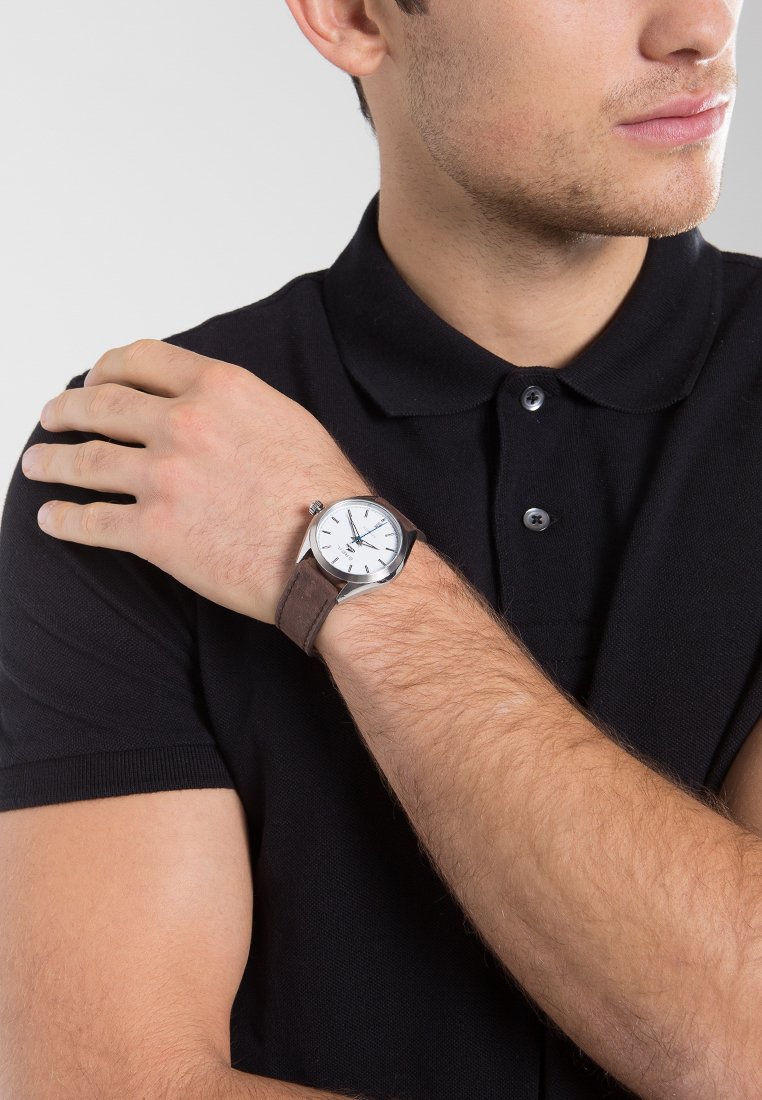 Reloj plateado con cara blanca, correa de cuero marrón y caja redonda, llevado en la muñeca de una persona con una camiseta polo negra.