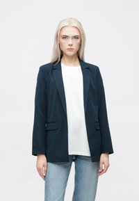 Jūrų laivyno švarkas su pritaikytu siluetu, apykakle ir dviem priekinėmis kišenėmis, uždėtas ant balto marškinio ir derintas su šviesiai mėlynomis džinsais.