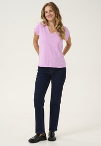 Rozā īsām piedurknēm V veida kakla T-krekls apvienots ar tumši zilām taisnām biksēm un melniem apaviem. Audumam ir strukturēta, viegla izskata virsma.