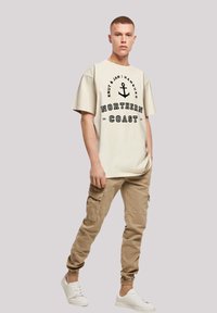 Cream oversized t-shirt with black graphic print and anchor design, paired with beige cargo pants featuring multiple pockets and elastic cuffs.