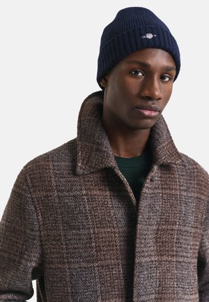 Giovane uomo che indossa un cappotto di lana marrone a quadri e un berretto lavorato a maglia blu navy con un piccolo logo ricamato, rivolto in avanti con un'espressione neutra.