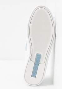 Bílá podešev tenisky s texturovaným povrchem, s modrou štítkovou značkou s nápisem "MICHAEL KORS." Hladké, zaoblené hrany a čistý design.