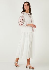 Blouse en lin blanc avec broderies florales rouges, associée à une longue jupe maxi blanche. Modèle portant des sandales argentées ornées.