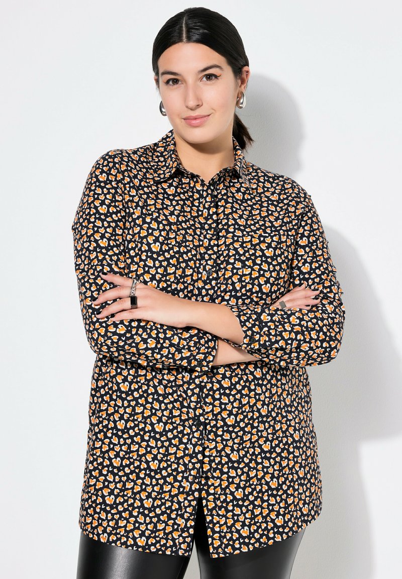 Sort skjorte med knapper og mønster af orange og creme farvede hjerter, lange ærmer og en afslappet pasform, parret med skinnende sorte leggings.