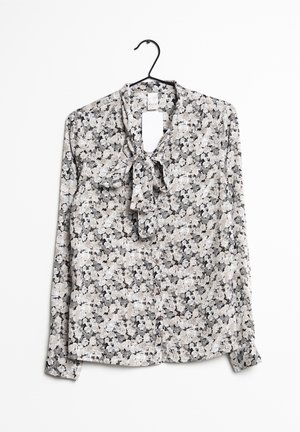 Langärmlige Bluse mit Blumenmuster in Schwarz, Weiß und Grau, mit Knopfleiste vorne und einer gebundenen Schleife am Ausschnitt an einem schwarzen Bügel.