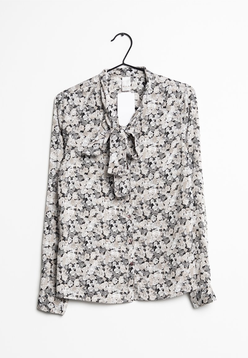 Blouse à manches longues à fleurs noires, blanches et grises, fermeture boutonnée devant et nœud noué au col sur un cintre noir.