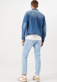 Chaqueta de mezclilla azul con un diseño desgastado, cierre de botones en la parte delantera y dos bolsillos laterales, combinada con pantalones vaqueros de lavado claro y zapatillas casuales.