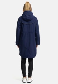 Marinblå vattentät parkas med huva, justerbart midjebälte och bakre ventil. Långa ärmar och något förlängd baksida. Tillsammans med svarta tajta jeans och vita sneakers.