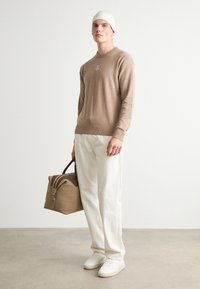 Μπεζ πουλόβερ με πλεκτές μανσέτες, κρεμ παντελόνι, λευκά αθλητικά παπούτσια και κρεμ πλεκτό σκουφάκι. Κρατάει μια καφέ τσάντα με σκούρους καφέ τόνους.