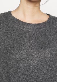 Pull en tricot gris avec un col rond. La texture semble douce et légèrement chinée, mettant en valeur une subtile variation de teintes.