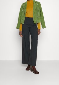 Grüne Wildleder-Bikerjacke, senfgelbes Langarmoberteil, schwarze weite Hose und glänzende bordeauxfarbene Stiefeletten mit Absatz. Glatte Texturen und figurbetonte Silhouetten.