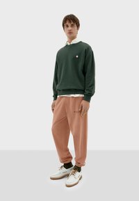 Felpa verde con un piccolo logo, indossata sopra una camicia bianca. Pantaloni jogger di colore pesco con una texture liscia, abbinati a scarpe bianche e calze verdi.