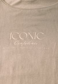 Světle béžové bavlněné tričko s kulatým výstřihem a potiskem v světle barvě: "ICONIC Confidence EST. 2026."
