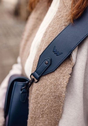 Marineblauer Leder-Schultergurt mit eingeprägtem Logo, befestigt an einer strukturierten braunen Tasche, mit einem metallischen Clip und einem weichen Stoffhintergrund.