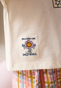 Bílá textilní etiketa s usměvavou květinovou postavičkou ukazující znaky míru a textem "SMILE EVERY DAY" a "SMILEYWORLD" na světle barevné látce.