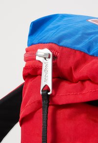 Sac à dos en tissu rouge et bleu avec une fermeture éclair argentée. Fabriqué dans un matériau durable, avec une surface texturée et une sangle noire.