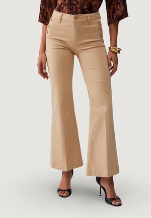 Donna con pantaloni beige a vita alta svasati con bottoni laterali, abbinati a tacchi neri aperti davanti e una maglia a maniche lunghe con motivo.