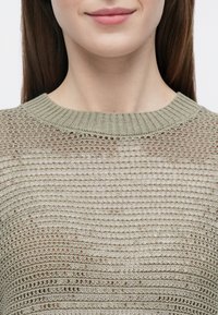 Pull en maille vert clair avec un col rond, présentant un motif à maille ouverte et un tissu texturé, des coutures visibles et des détails côtelés.