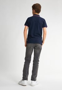 Námornícké polo tričko s červeným lemováním, nošené s šedými džíny a bílými teniskami. Pohled ukazuje zadní část outfitu, který má krátké rukávy a volný střih.