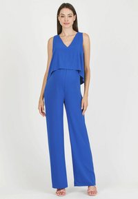 Mono azul royal con escote en V y parte superior superpuesta, ajuste holgado, piernas anchas y textura suave; combinado con sandalias de tiras rosas claras.