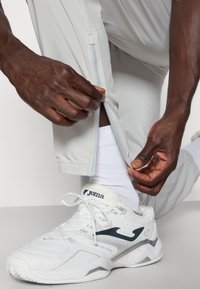 Lichtgrijze sportbroeken met een zijrits, gecombineerd met witte sneakers met marineblauwe accenten en een gestructureerd oppervlak.