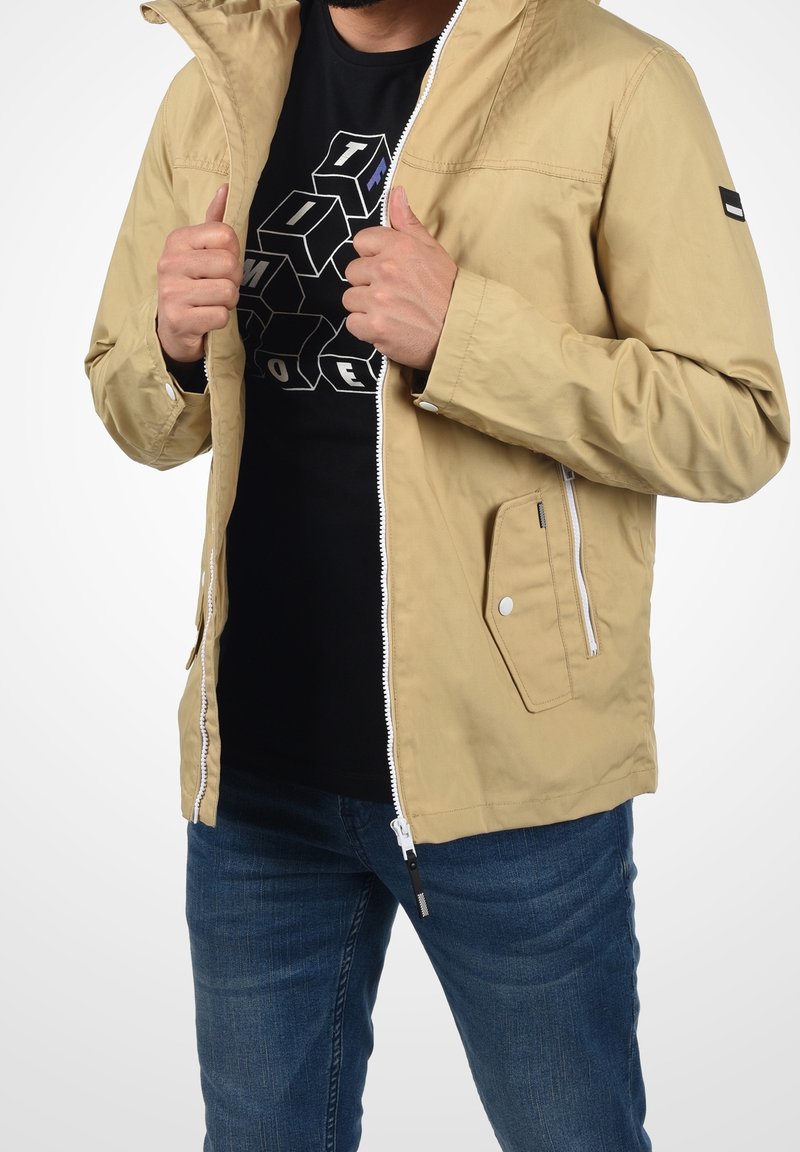 Homme portant une veste beige zippée sur un t-shirt noir graphique et un jean bleu, tenant la veste ouverte avec les deux mains.