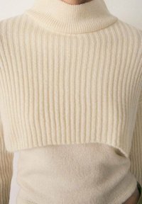 Pull tricoté de couleur crème avec un col montant, présentant une texture côtelée et une coupe courte. Tissu doux et lisse avec une coupe ajustée.