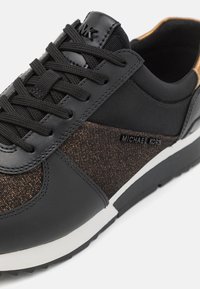 Baskets noires et marron avec lacets noirs, panneau latéral texturé, bord de semelle blanc et logo Michael Kors sur le côté et la languette.