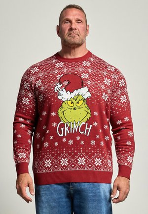 Hombre llevando un suéter rojo de Navidad con copos de nieve blancos y una gran cara del Grinch con un gorro de Santa en el centro.
