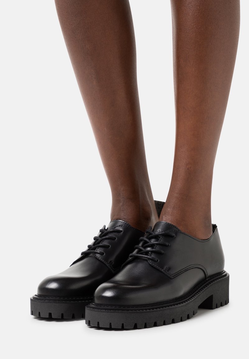 Chaussures en cuir noires épaisses à lacets portées sur des pieds aux chevilles visibles, dotées de semelles épaisses à relief et d'une finition lisse et polie.