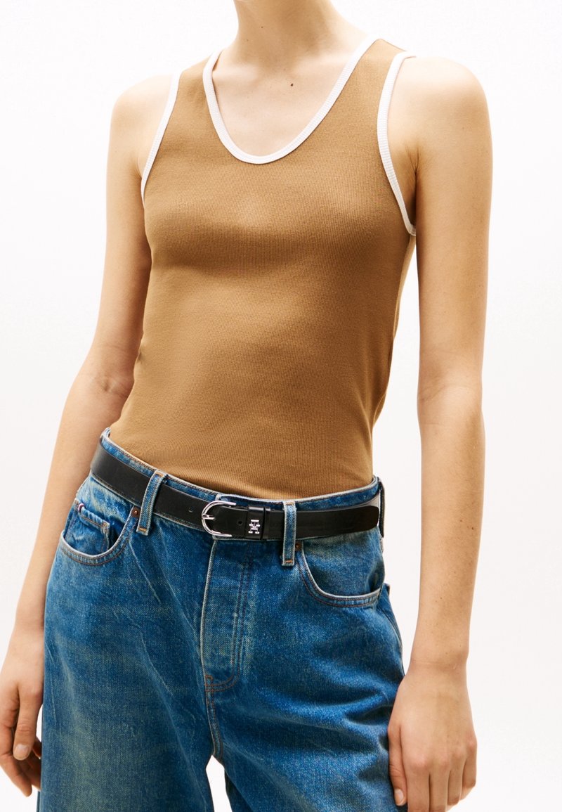 Personne portant un haut marron sans manches avec un bord blanc et un jean bleu taille haute maintenu par une ceinture noire avec une boucle argentée.