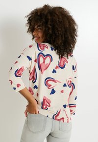 Un pull beige clair avec des motifs de cœurs multicolores en rose et marine. Il a des manches trois-quarts et une coupe décontractée.
