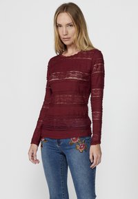 Top lungo borgogna a maniche lunghe con dettagli in pizzo, tessuto texture, scollatura a onde e design aderente, abbinato a jeans blu ricamati.
