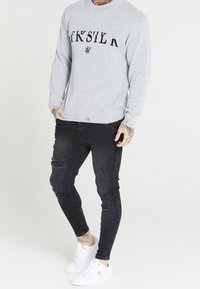 Pull en maille gris avec logo noir, jeans slim noirs avec des détails déchirés, et baskets blanches avec des accents dorés.