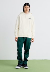 Sudadera crema con bolsillo frontal, que presenta un pequeño logo; combinada con pantalones deportivos verde oscuro y zapatillas negras. Textura de tela suave.
