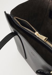 Sac à main en cuir noir avec fermeture éclair dorée partiellement ouverte, montrant l'intérieur en daim foncé et deux poignées sur fond blanc.