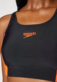 Top corto deportivo negro con acentos naranjas y logo de Speedo. Presenta una textura suave y un escote redondeado para un diseño ajustado.