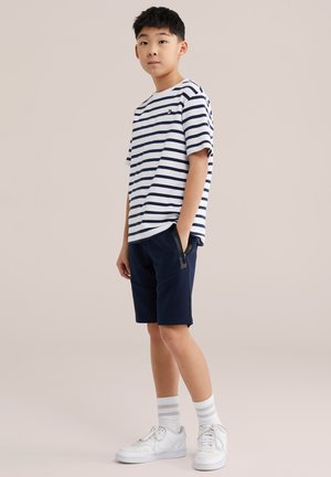 Jeune garçon debout, les mains dans les poches, portant un t-shirt blanc à rayures bleues marine, un short bleu marine, des chaussettes blanches et des baskets blanches sur un fond beige.