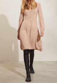 Ljusrosa stickad klänning med långa, puffiga ärmar och en scallopslagd fåll som har geometrisk broderi längs botten. Kombineras med svarta höga stövlar.