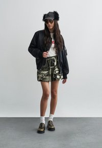 Schwarze Bomberjacke, weißes T-Shirt mit rotem Logo, Tarnshorts, weiße Socken und klobige bronzefarbene Schuhe. Schwarze Kappe mit Pelzakzent und Sonnenbrille.