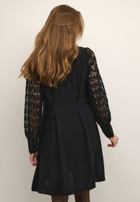 Black dress featuring lace long sleeves, a fitted bodice, and a flared skirt with pleats. Smooth fabric with a matte finish.