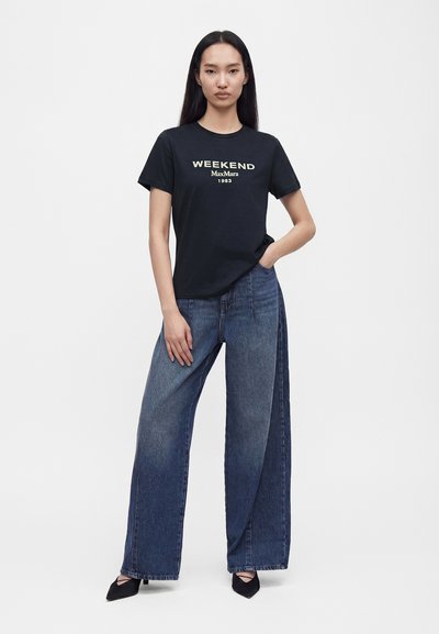 Femme debout, une main dans la poche, portant un t-shirt noir "Weekend Max Mara", un jean large bleu et des chaussures noires à bout pointu.