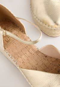 Guldig espadrille med texturerad ovandel, korksula och justerbar ankelrem. Har sydd kant och öppen tådesign.