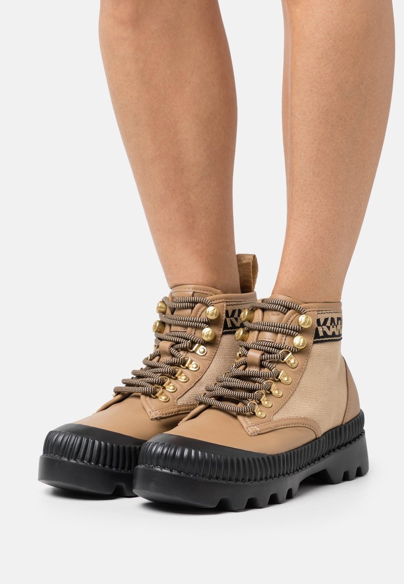 Bottes de randonnée montantes beige et marron clair avec semelles noires épaisses, œillets dorés et lacets à motifs portées sur des jambes nues.