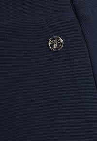 Tessuto di colore blu navy con texture, caratterizzato da un piccolo emblema rotondo in metallo dorato con un design intagliato complesso, attaccato da due cuciture.