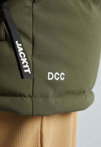 Zelená prošívaná tkanina s bílým výšivkou "DCC" a černým taháčkem zipu označeným "JACK11", na bežovém texturovaném materiálu pod ním.