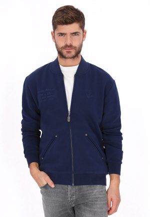 Jonge man met kort bruin haar en baard, gekleed in een marineblauw jack met rits en grijze spijkerbroek, staand met één hand in zijn zak tegen een witte achtergrond.