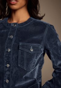 Granatowa kurtka dżinsowa o gładkiej fakturze, z metalowymi guzikami, kieszenią na piersi oraz detalami w kontrastowych szwach.
