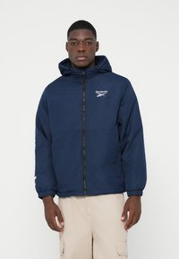 Tânăr îmbrăcat cu o jachetă cu glugă Reebok de culoare bleumarin, cu fermoar în față, și pantaloni cargo bej, stând în fața unui fundal alb simplu.